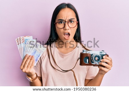 Young asian woman holding vintage camera and swedish krona in shock face, looking skeptical and sarcastic, surprised with open mouth 
