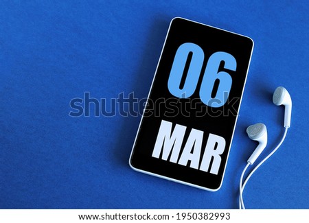 March 06. 6 st day of the month, calendar date. Smartphone and white headphones on a blue background. Place for your text. Springtime month, day of the year concept.