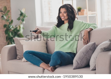Full body photo of young afro woman happy positive smile sit sofa rest watch tv switch channel remote controller indoors