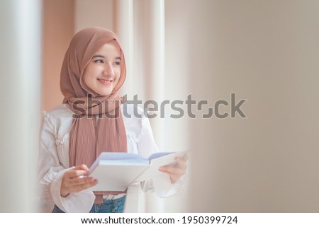 A hijab teenager is reading a book and smiling when greeted Royalty-Free Stock Photo #1950399724