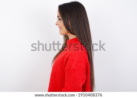 Profile of smiling young beautiful Caucasian woman wearing red knitted sweater over white studio wall with healthy skin, has contemplative expression, ready to have outdoor walk.