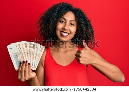 Beautiful african american woman with afro hair holding 100 danish krone banknotes smiling happy and positive, thumb up doing excellent and approval sign 