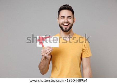 Young caucasian smiling bearded attractive handsome happy man 20s wearing casual yellow basic t-shirt hold gift voucher flyer mock up looking camera isolated on grey color background studio portrait