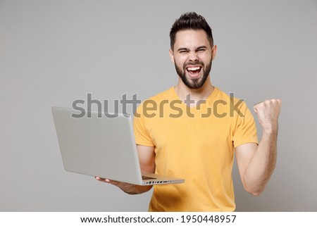 Young caucasian smiling bearded happy copywriter freelancer man 20s wearing casual yellow basic t-shirt hold laptop pc computer show thumb up gesture isolated on grey color background studio portrait