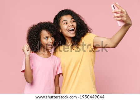 Happy african american young woman little kid girl sisters in t-shirts hugging doing selfie shot on mobile phone doing winner gesture isolated on pink background studio portrait. Family day concept