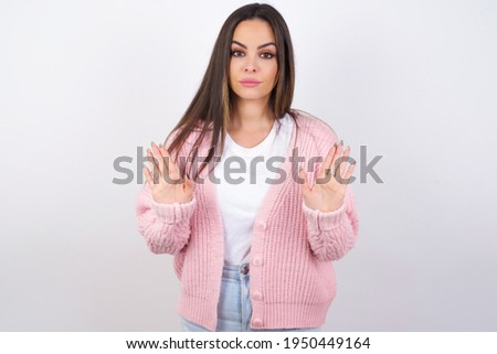 Serious Young beautiful caucasian woman wearing pink knitted jacket over white wall pulls palms towards camera, makes stop gesture, asks to control your emotions and not be nervous