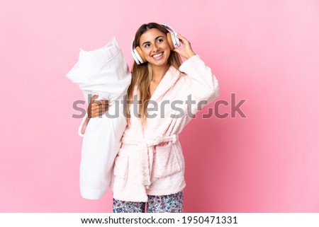 Young hispanic woman over isolated pink background in pajamas and holding a pillow and listening music