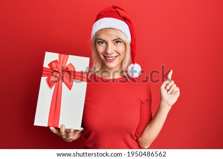 Young blonde girl wearing christmas hat and holding a gift smiling happy pointing with hand and finger to the side 