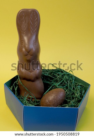 An Easter chocolate bunny and a chocolate egg in a blue box with green pieces of paper lie on a yellow background side view . Easter holiday. milk chocolate rabbit