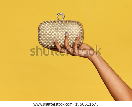Hand of a young black woman holding a stylish gold metallic pochette, purse or small handbag on display over a yellow studio background with copyspace Royalty-Free Stock Photo #1950511675