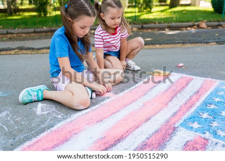 Side view of girls drawing American flag with colored chalks on the sidewalk near the house on sunny summer day. Kids painting outside. Creative development of children.  Independence Day concept