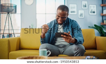 Happy Black African American Man Playing Video Game on Smartphone App and Win, Showing YES Gesture. He is Sitting on a Sofa and Resting in Living Room, Having Fun Over the Internet. Royalty-Free Stock Photo #1950543853