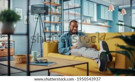 Handsome Black African American Man Working on Laptop Computer while Lying on a Sofa in Cozy Living Room. Freelancer Working From Home. Browsing Internet, Using Social Networks, Having Fun in Flat. Royalty-Free Stock Photo #1950543937