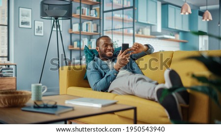 Excited Black African American Man Using Smartphone while Resting on a Sofa in Living Room. Happy Man Smiling at Home and Chatting to Colleagues and Clients Over the Internet. Using Social Networks. Royalty-Free Stock Photo #1950543940
