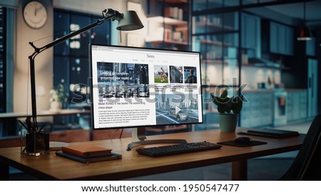 Shot of a Desktop Computer with Latest News Web Page Showing On Screen Standing on the Wooden Desk in the Creative Cozy Living Room. In the Background Warm Evening Lighting and Open Space Studio. Royalty-Free Stock Photo #1950547477