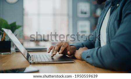 Close Up on Hands of a Black African American Man Working on Laptop Computer while Sitting Behind Desk in Cozy Living Room. Freelancer Working From Home. Browsing Internet, Using Social Network. Royalty-Free Stock Photo #1950559639