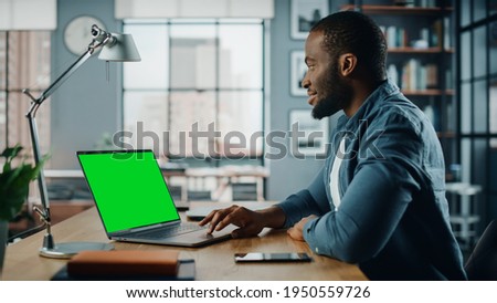 Handsome Black African American Specialist Working on Laptop Computer with Green Screen Mock Up Display at Home Living Room. Freelance Man Chatting to Clients Over the Internet on Social Networks.
