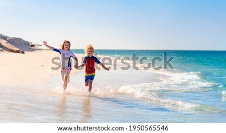 Kids playing on tropical beach. Children swim and play at sea on summer family vacation. Sand and water fun, sun protection for young child. Little boy and girl running and jumping at ocean shore.