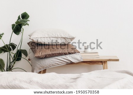 Trendy Scandinavian interior still life. Velvet and linen cushions on vintage wooden bench, table. Cup of coffee on pile of books and monstera potted plant. White wall background. Blurred bed Royalty-Free Stock Photo #1950570358