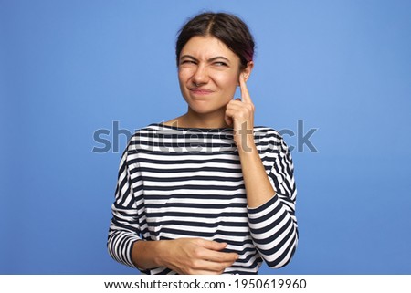 Horizontal shot of frustrated young Caucasian female with facial piercing holding finger at her ear, trying to remove wax, having painful grimace, frowning, posing isolated against blue background Royalty-Free Stock Photo #1950619960