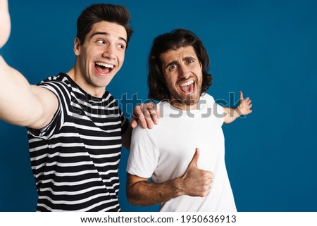White excited two men gesturing thumb up while taking selfie photo isolated over blue background