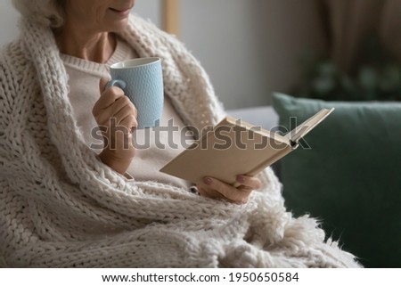 Old retired 70s lady reading book at home, drinking hot tea, resting on sofa in cozy living room. Mature reader wrapped in knitted blanket relaxing with novel over cup of coffee or chocolate. Close up