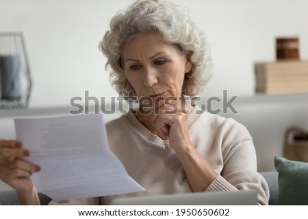 Focused mature 60s retired lady receiving letter and reading document at home. Serious middle aged woman checking bank loan notification, tax form, making online payment of bill or invoice Royalty-Free Stock Photo #1950650602