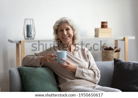 Portrait of happy senior 60s woman holding cup, sitting on couch, looking at camera. smiling. Female pensioner relaxing on sofa at home, drinking hot tea or coffee, speaking during video call Royalty-Free Stock Photo #1950650617