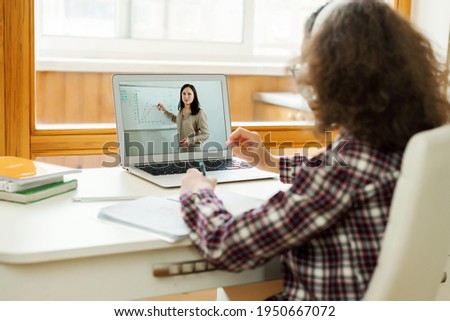 Teen girl is doing her homework at desk indoors. Famale teenager watching video at desk study distant form home using laptop. Online learning