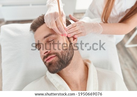 Concept of cosmetology and facial. A woman beautician makes face and beard modeling for a man waxing epilation. Depilation with wax Royalty-Free Stock Photo #1950669076
