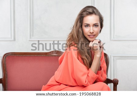 Portrait of nice young dark blonde woman in pink dress in classic interior on red sofa