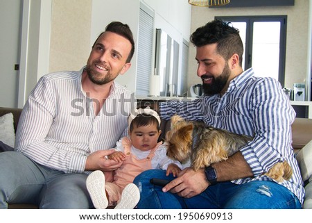 gay couple with their young daughter and dog sitting on the sofa watching television. The girl watches the cartoons attentively