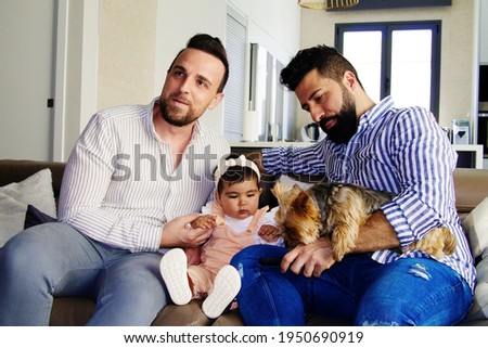 gay couple with their young daughter and dog sitting on the sofa watching television. The girl watches the cartoons attentively.
