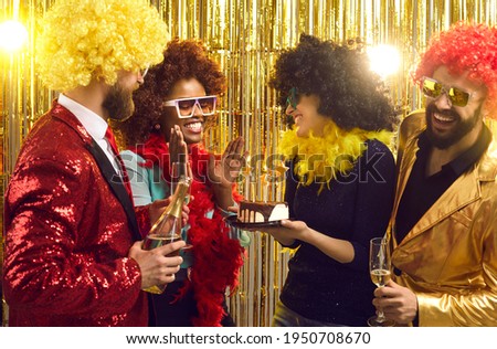 Group of happy friends in retro outfits, feather boas and Afro hair wigs with cake and champagne congratulating young black woman at 70s disco music nightclub fancy dress fashion themed birthday party Royalty-Free Stock Photo #1950708670