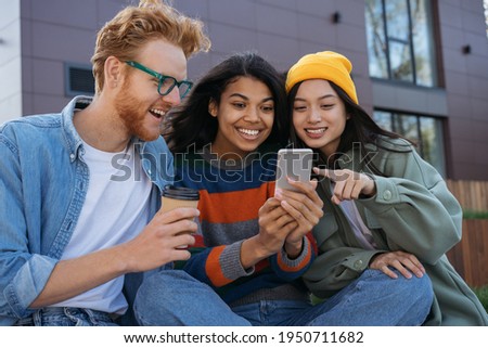 Group of smiling multiracial friends using mobile phone, watching video, shopping online, having fun outdoors.  Young happy students studying, learning language, communication together  Royalty-Free Stock Photo #1950711682