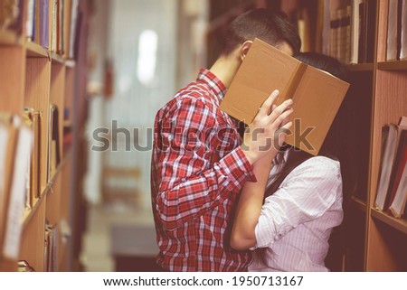 Photo of man and woman kissing in library and close their face by book. Young couple of boy and girl hiding for spending time together. Students
