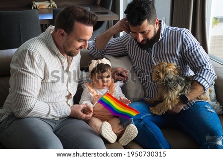 gay couple with their young daughter and dog sitting on the sofa watching television. The little girl watches the cartoons attentively and holds the gay pride flag in her hand