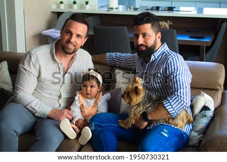 gay couple with their young daughter and dog sitting on the sofa watching television. The girl watches the cartoons attentively