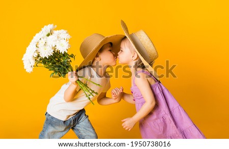 portrait of a little girl and a boy in straw hats kissing on a yellow background with space for text. A boy holds a bouquet of white flowers