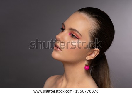 High beauty photo of a lovely young girl with blue eyes, round pink earrings and wonderful professional makeup, with long brown hair. Posing over gray background. Close-up. Studio shot. Portrait