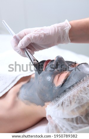 Cosmetologist uses brush to apply cosmetic mask to woman's face.Woman's face is covered with an alginate mask. Cosmetic procedure. A gray, frozen mass on woman's face.Vertical photo.Concept of beauty.