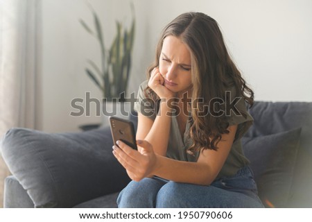 Bored young woman spends leisure time in social networks, holds smartphone. Depressed melancholy female with a mobile phone sitting on the couch at home