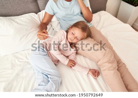 A picture of a mother checking her daughters temperature and calls a doctor , girl is having fever. Both laying on the bed with a plant on a background