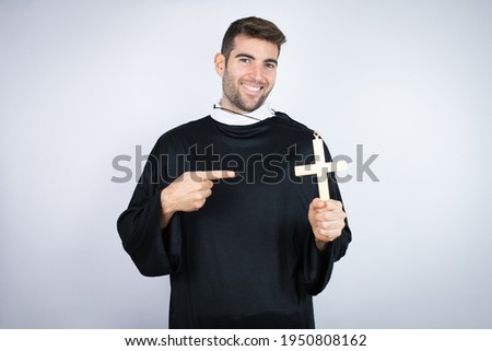 Young hispanic man wearing priest uniform standing over white background holding crucifix smiling happy pointing with hand and finger