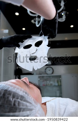 The beautician holds a fabric moisturizing face mask. A man makes facial masks in a cosmetic clinic. Men's cosmetology. Youth preservation and skin care concept. Vertical photo. Bottom view.