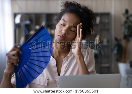 Unwell young African American woman use waver feel overheated work on computer at home office. Tired biracial female wave with hand fan breathe fresh air, suffer from conditioner lack indoors. Royalty-Free Stock Photo #1950865501