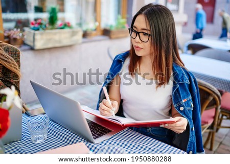 Young ethnic female student with eyeglasses sitting at table reading information on laptop and taking notes in notepad doing homework