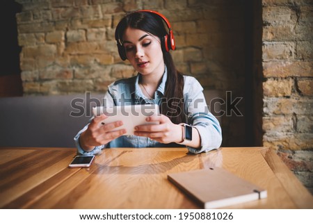Young wistful female in casual clothes sitting at wooden table while listening to music in wireless headphones and surfing tablet during break