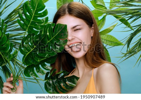 cheerful woman shows tongue and stands near green leaves of palm tree on blue background