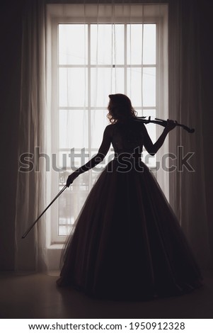 Pretty slender young woman with violin at window. Female in stage image in sunlight. Emotional girl in dress standing at window in interior room. Actress show. Theatre performance concept. Copy space